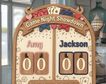 May include: A wooden game night scoreboard with the words "Game Night Showdown". It features spaces for two players, dice, hearts, playing cards, and game controllers. The names "Amy" and "Jackson" are displayed above the score boxes.