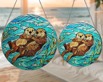 May include: Two round stained glass panels depicting otters in turquoise water with bubbles, fish, and seaweed. The otters are brown and tan, hugging each other. Each panel is suspended by a silver chain. The background shows a beach scene.