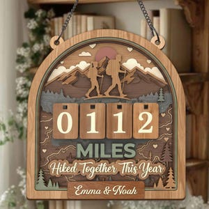 Op de afbeelding: Houten bord met wandelthema met een berglandsilhouet, wandelaars en de tekst "0112 MILES Hiked Together This Year Emma & Noah". Het bord is bruin, met een ketting om op te hangen.
