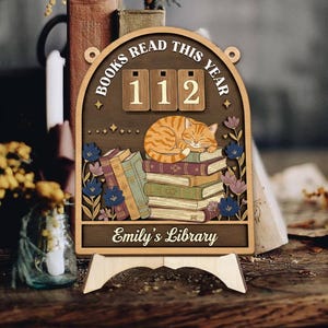 May include: Wooden bookshelf decor with the text "BOOKS READ THIS YEAR" and the number "112". A sleeping orange tabby cat rests on a stack of books. The bottom of the decor reads "Emily's Library".