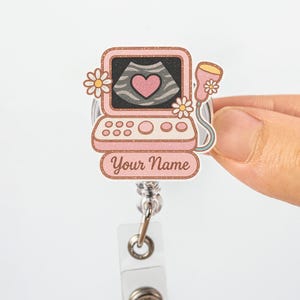 May include: A pink and white ultrasound badge reel with a floral design. The badge features a heart-shaped image on the screen and the text "Your Name". The reel is attached to a clear plastic holder.