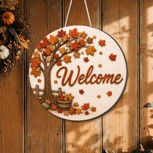 May include: A circular wooden sign featuring a printed tree with orange and red autumn leaves, a wooden fence, and a barrel. The word Welcome is written in a brown script font in the center. The sign hangs from a white rope against a wood background.