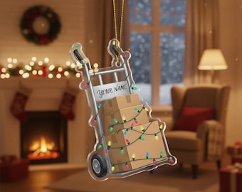 Op de afbeelding: Transparante acryl kerstversiering in de vorm van een steekwagen beladen met kartonnen dozen. De steekwagen heeft een bordje met "Your Name" en is versierd met kleurrijke kerstverlichting. De versiering hangt aan een gouden touwtje.