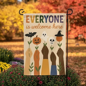 Pode incluir: Uma bandeira de jardim com o texto "EVERYONE is welcome here" em letras coloridas. A bandeira apresenta ilustrações de mãos segurando flores temáticas de Halloween, incluindo um morcego, abóbora, fantasma, caveira e chapéu de bruxa. A bandeira é exibida ao ar livre.