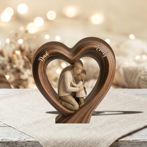 May include: A wooden heart-shaped sculpture with a father and daughter embracing inside. The sculpture is brown with the words "Dad" and "Emily" inscribed on the heart. The figures are in neutral tones.