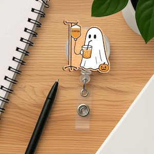May include: A Halloween-themed badge reel with a cartoon ghost drinking from a cup, a pumpkin, and an IV drip. The IV bag reads "PUMPKIN". The reel has a clear plastic strap and a metal clip.