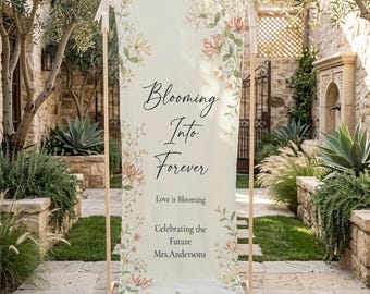 Può includere: Un cartello nuziale color crema con la scritta "Blooming Into Forever" e decorazioni floreali. Il cartello è esposto su una cornice di legno e include il testo "Love is Blooming" e "Celebrating the Future Mrs. Anderson".