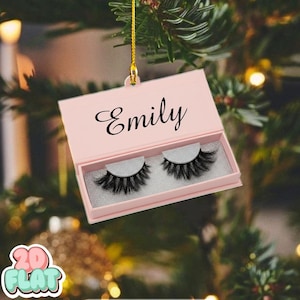 May include: A pink box-shaped Christmas ornament with the name "Emily" in black script. Inside the box are two sets of black false eyelashes. The ornament hangs from a gold string against a blurred background of Christmas lights and greenery.