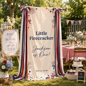 May include: An outdoor party scene with a banner reading "Little Firecracker Jackson is One!" and "July 4, 2026." The banner is decorated with red, white, and blue ribbons and stars. A sign says "Baby in Bloom - this - 4th of July."