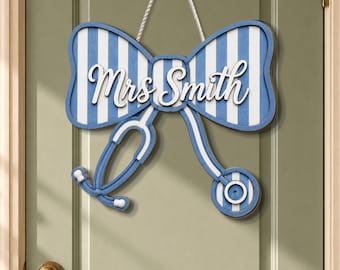 Puede incluir: Un letrero de puerta de madera con forma de lazo con rayas azules y blancas, un estetoscopio blanco y el nombre "Mrs Smith" en escritura blanca. El letrero está colgado en una puerta.
