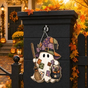 May include: A hanging wall sign shaped like a ghost wearing a patchwork witch hat. The ghost holds a broom and a black cauldron with a star, featuring various colorful fabric-style patches on its body and hat, suspended by a white rope.