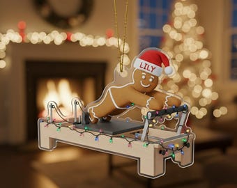Könnte beinhalten: Ein klarer Weihnachtsbaumschmuck aus Acryl in Form eines Lebkuchenmanns mit Weihnachtsmütze und dem Namen "LILY" darauf, der auf einem Pilates-Reformer trainiert. Der Schmuck ist mit bunten Lichtern und dem Text "2D FLAT" verziert.