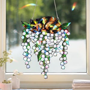 Pode incluir: Um apanhador de sol de gato em vitral, apresentando um gato dormindo com marcas marrons, pretas e amarelas, descansando sobre folhas verdes e flores brancas de lírio-do-vale. O apanhador de sol pende de uma corrente prateada, criando um adorno decorativo para janelas.