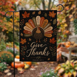 Op de afbeelding: Een zwarte, rechthoekige tuin vlag met een kalkoen in het midden met een kleurrijke, gedessineerde staartwaaier. De tekst "Give Thanks" en "PSALM 107:1" is in gouden letters gedrukt. Het ontwerp bevat verspreide herfstbladeren en takjes.