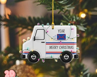 Op de afbeelding: Een witte kerstversiering in de vorm van een postwagen met rode en blauwe strepen en de woorden "MERRY CHRISTMAS" en "YEAR". De versiering is versierd met een slinger van gekleurde lichtjes en een gouden draad om op te hangen.
