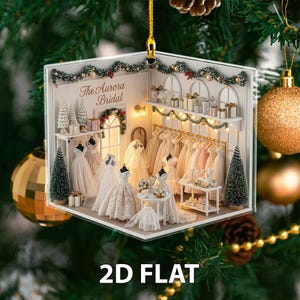 Könnte beinhalten: Ein 2D-flacher Weihnachtsschmuck mit einer Brautmodenszene. Der Schmuck zeigt Brautkleider auf Schaufensterpuppen und einer Kleiderstange, mit dem Text "The Aurora Bridal". Der Schmuck ist mit Miniatur-Weihnachtsbäumen und Girlanden verziert.