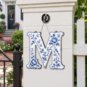 May include: A decorative letter M wall hanging featuring a white base with a blue floral pattern, including detailed illustrations of blooming peonies and a small bird perched on a branch, suspended by a thin white cord from a black wall hook.
