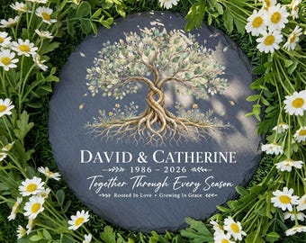 Può includere: Una pietra rotonda, grigio scuro, con un disegno di albero e i nomi "DAVID & CATHERINE" e le date 1986-2026. È presente anche il testo "Together Through Every Season" e "Rooted In Love • Growing In Grace". La pietra è circondata da margherite bianche e verde.