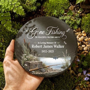 May include: A round, dark gray slate memorial stone with the words "Gone Fishing" and "In Loving Memory Of Robert James Walker 1952-2025". The stone features a fishing scene with a tackle box, fishing rod, and lake.