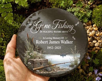 Può includere: Una lapide rotonda in ardesia grigio scuro con le parole "Gone Fishing" e "In Loving Memory Of Robert James Walker 1952-2025". La pietra presenta una scena di pesca con una scatola da pesca, canna da pesca e lago.