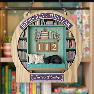 May include: Wooden bookshelf-shaped wall decor with the text "BOOKS READ THIS YEAR" and "Lucie's Library". The interior features a miniature library scene with a sleeping black cat, pillows, and the number "112".