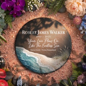 May include: A circular, dark grey memorial stone with the name "ROBERT JAMES WALKER" and dates "1953 - 2025" in white text. The stone features a beach scene with the text "Your Love Flows On Like The Endless Sea". Surrounded by flowers.
