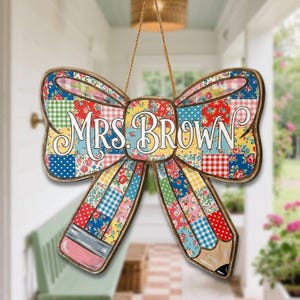 May include: A wooden bow-shaped sign with a pencil design, featuring the text "MRS. BROWN". The sign is decorated with a patchwork pattern of floral and gingham fabrics in various colors, and is suspended by a natural rope.