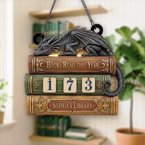 May include: A decorative sign featuring a sleeping dragon atop three stacked books. The top book reads "Books Read This Year", the middle book displays "173", and the bottom book says "Sophia's Library". The sign is suspended by a chain.