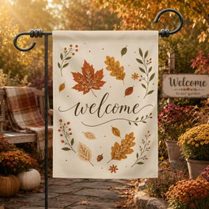 Pode incluir: Uma bandeira de jardim na cor creme com a palavra welcome em letra cursiva castanho-escura, rodeada por um padrão de folhas de outono laranja, amarelas e castanhas, pequenos ramos e uma bolota, suspensa num suporte de metal preto.