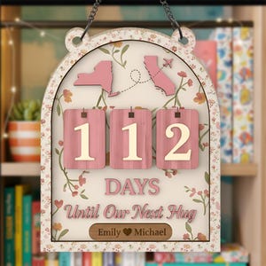 May include: A wooden countdown sign with a floral design and the text "112 DAYS Until Our Next Hug." The sign features pink cutouts of New York and California, with a small airplane. The names "Emily" and "Michael" are at the bottom.