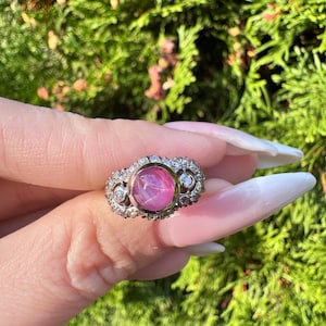 Op de afbeelding: Een vintage ring met een grote, ronde, roze edelsteen in het midden, omringd door kleinere, heldere edelstenen. De band van de ring is versierd, met gedetailleerd metaalwerk. De ring wordt tegen een achtergrond van groen gebladerte gehouden.