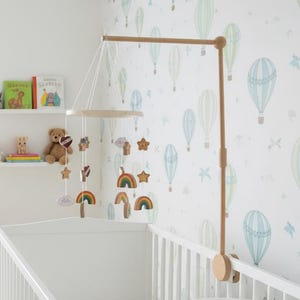 May include: A white crib in a nursery setting, with a wooden mobile featuring rainbows, stars, and clouds. The room has a light color scheme, with a rocking chair and a shelf with books and stuffed animals. The wallpaper has a hot air balloon pattern.