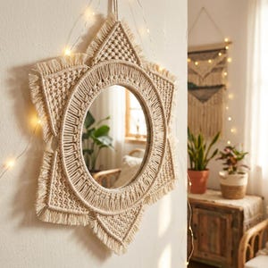 May include: A star-shaped macrame mirror with a circular mirror in the center. The off-white macrame has a fringed edge and is hanging on a white wall. Decorative lights are strung around the mirror.