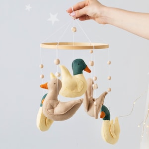 Pode incluir: Móvel de bebê com patos de feltro em tons de bege, amarelo e verde, suspensos em um anel de madeira. Os patos têm bicos laranja e são presos com cordas e contas de madeira. Uma mão segura o móvel.