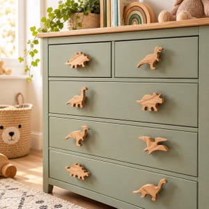 May include: A sage green wooden dresser featuring six drawers, each equipped with a unique light-colored wooden dinosaur-shaped knob. The dresser has a natural wood top surface and a clean, rectangular frame design with smooth drawer fronts.