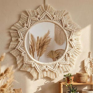 May include: A round, cream-colored macrame mirror with a sunburst design. The mirror's frame features intricate knotting and fringe details. The mirror is hanging on a neutral-toned wall, reflecting dried plants and other decor.