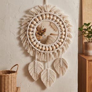 May include: A round, cream-colored macrame mirror with a fringe and wooden bead design. Three cream-colored, leaf-shaped macrame pieces hang below the mirror. The mirror is hanging on a light-colored wall.