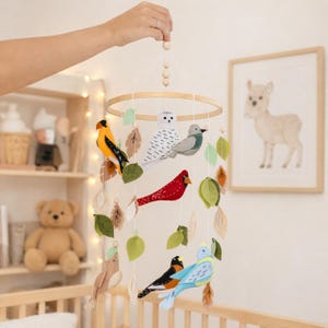 May include: A handmade baby mobile with felt birds in colours such as yellow, red, blue, and grey, hanging from a wooden hoop. The mobile also features felt leaves in green and brown, creating a whimsical nursery decoration.
