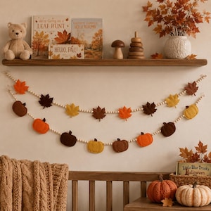 May include: Two decorative garlands featuring felt pumpkins and maple leaves in shades of orange, yellow, and brown, strung on light-coloured wooden beads. The garlands are displayed against a wall, hanging in a draped, layered arrangement.
