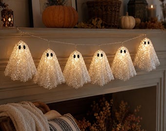 May include: A string of six illuminated ghost-shaped decorations hanging in a row. Each ghost is crafted from a textured, translucent white material with two small black oval eyes and contains a warm glowing light inside the hollow body.