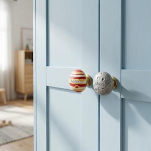 May include: Two round cabinet knobs attached to a light blue door. One knob features a painted Jupiter design with red and tan bands and the text Jupiter. The other knob is gray with a cratered surface and the text Moon. Both have gold bases.