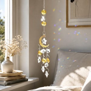 May include: A hanging suncatcher featuring a gold-toned crescent moon charm and a five-pointed star, accented with multiple clear and yellow faceted crystal beads suspended on a silver-coloured metal chain, creating light refractions.