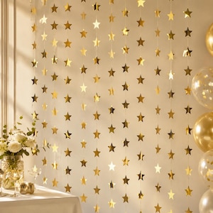 Op de afbeelding: Een verticale slingerachtergrond met talloze metallic gouden stervormige uitsparingen opgehangen aan dunne touwtjes. De sterren zijn gerangschikt in meerdere parallelle rijen, wat zorgt voor een glinsterende, reflecterende weergave tegen een muur.