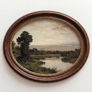 Könnte beinhalten: Ein ovales Landschaftsgemälde in einem dunkelbraunen Holzrahmen mit goldener Innenkante. Das Kunstwerk zeigt einen ruhigen Fluss, der durch ein grasbewachsenes Feld mit Bäumen unter einem weichen, bewölkten Abendhimmel fließt, in gedeckten Grün- und Brauntönen.