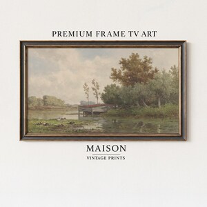 Könnte beinhalten: Ein rechteckiges Landschaftsgemälde in einem dunkelbronzenen Rahmen mit der Aufschrift PREMIUM FRAME TV ART und MAISON VINTAGE PRINTS. Das Bild zeigt eine ruhige Flusslandschaft mit einem kleinen Holzbau, grünen Bäumen und bewölktem Himmel.