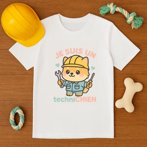Peut inclure: T-shirt blanc avec un dessin de chien de dessin animé portant un casque jaune et tenant des outils. Le texte "JE SUIS UN techniCHIEN" est imprimé sur le t-shirt en rose et bleu. Un casque jaune, un jouet pour chien et un jouet en forme d'os sont également visibles.