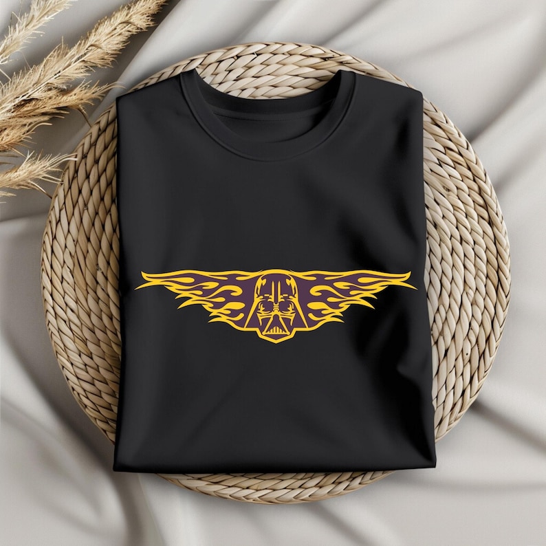 May include: Black t-shirt featuring a yellow and purple graphic of Darth Vader's helmet with flame-like wings. The design is centered on the shirt. The shirt is folded and placed on a woven, circular surface.
