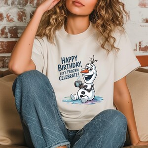 May include: Cream-colored t-shirt featuring a graphic of Olaf from Frozen, holding a mug. The text "HAPPY BIRTHDAY, LET'S FROZEN CELEBRATE!" is printed in blue. The shirt is a short-sleeved crew neck.