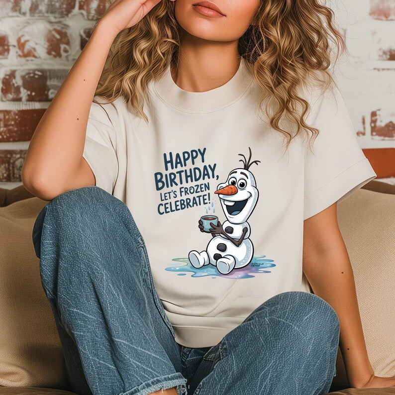 May include: Cream-colored t-shirt featuring a graphic of Olaf from Frozen, holding a mug. The text "HAPPY BIRTHDAY, LET'S FROZEN CELEBRATE!" is printed in blue. The shirt is a short-sleeved crew neck.