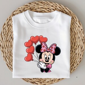 May include: White sweatshirt featuring a cartoon Minnie Mouse holding red heart-shaped balloons. Minnie Mouse is wearing a pink dress and a pink bow with heart patterns. The sweatshirt is folded on a woven surface.